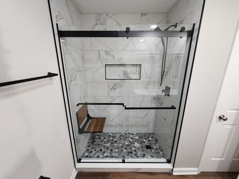 Modern shower with marble tile, glass doors, black hardware, and a wooden seat.