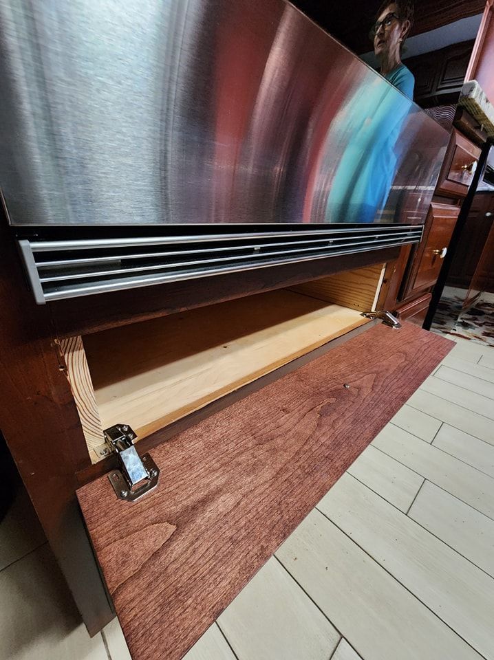 Open cabinet door beneath a stainless steel appliance, showing a wooden interior and person in background.