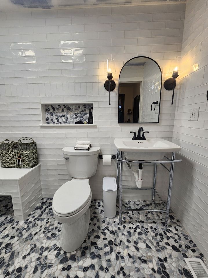 Bathroom with white brick tile walls, pebble tile floor, toilet, sink, and arched mirror.