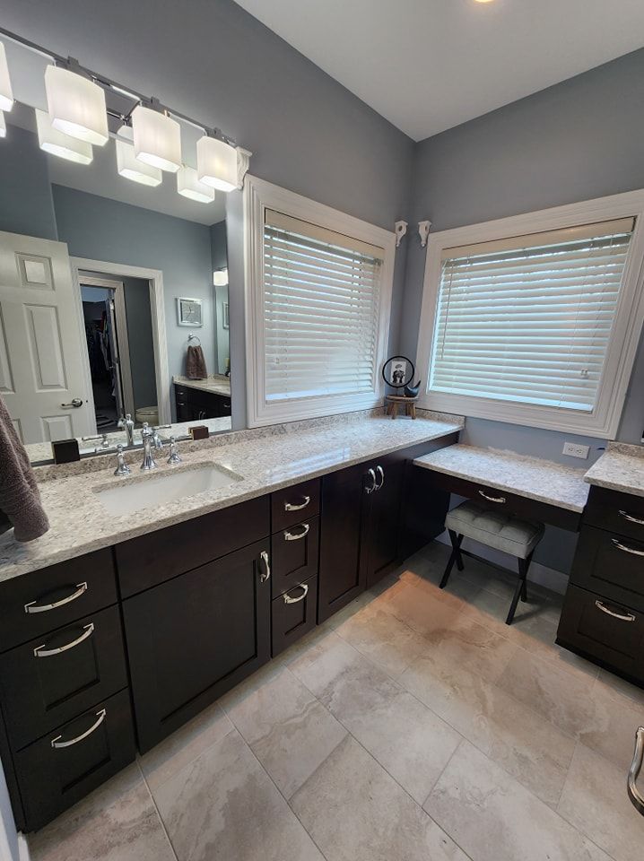 Bathroom with dark brown vanity, granite countertop, two windows, and light gray walls.