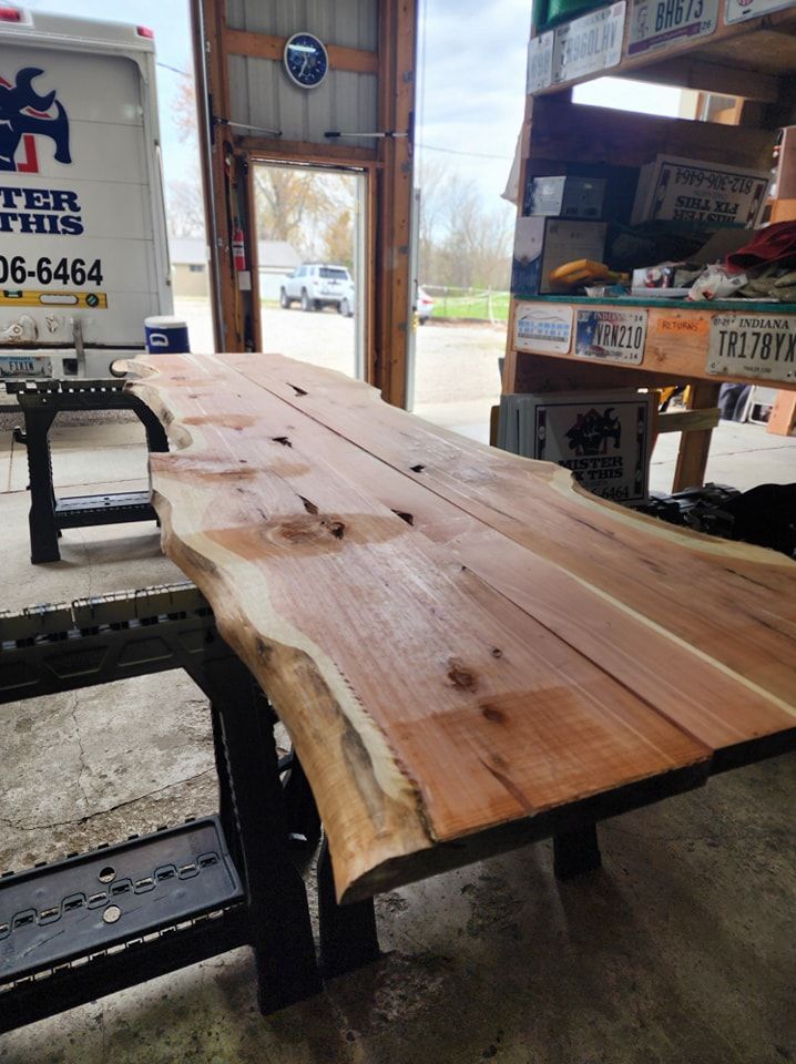 Live-edge wooden slab on black sawhorses inside a workshop. Door open to outside.