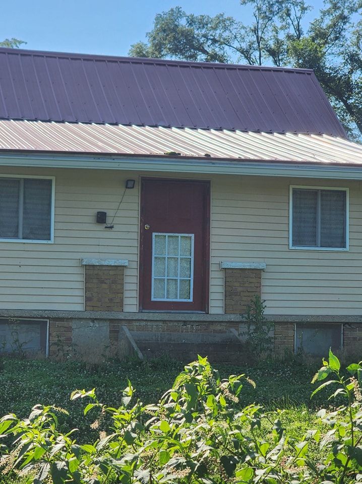 Beige house with burgundy door and roof, two windows, and overgrown vegetation.