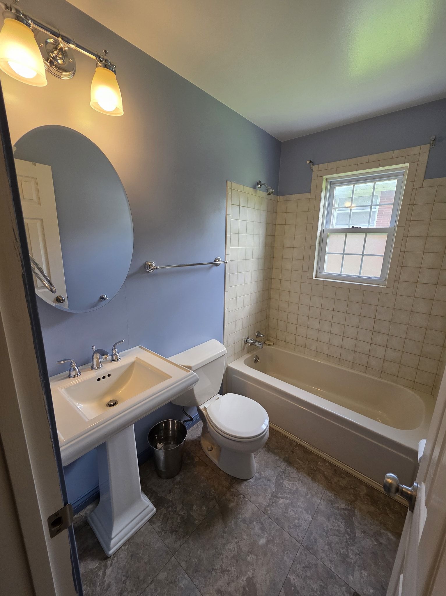 Bathroom with blue walls, white sink and toilet, a tub with a tiled surround, and a small window.