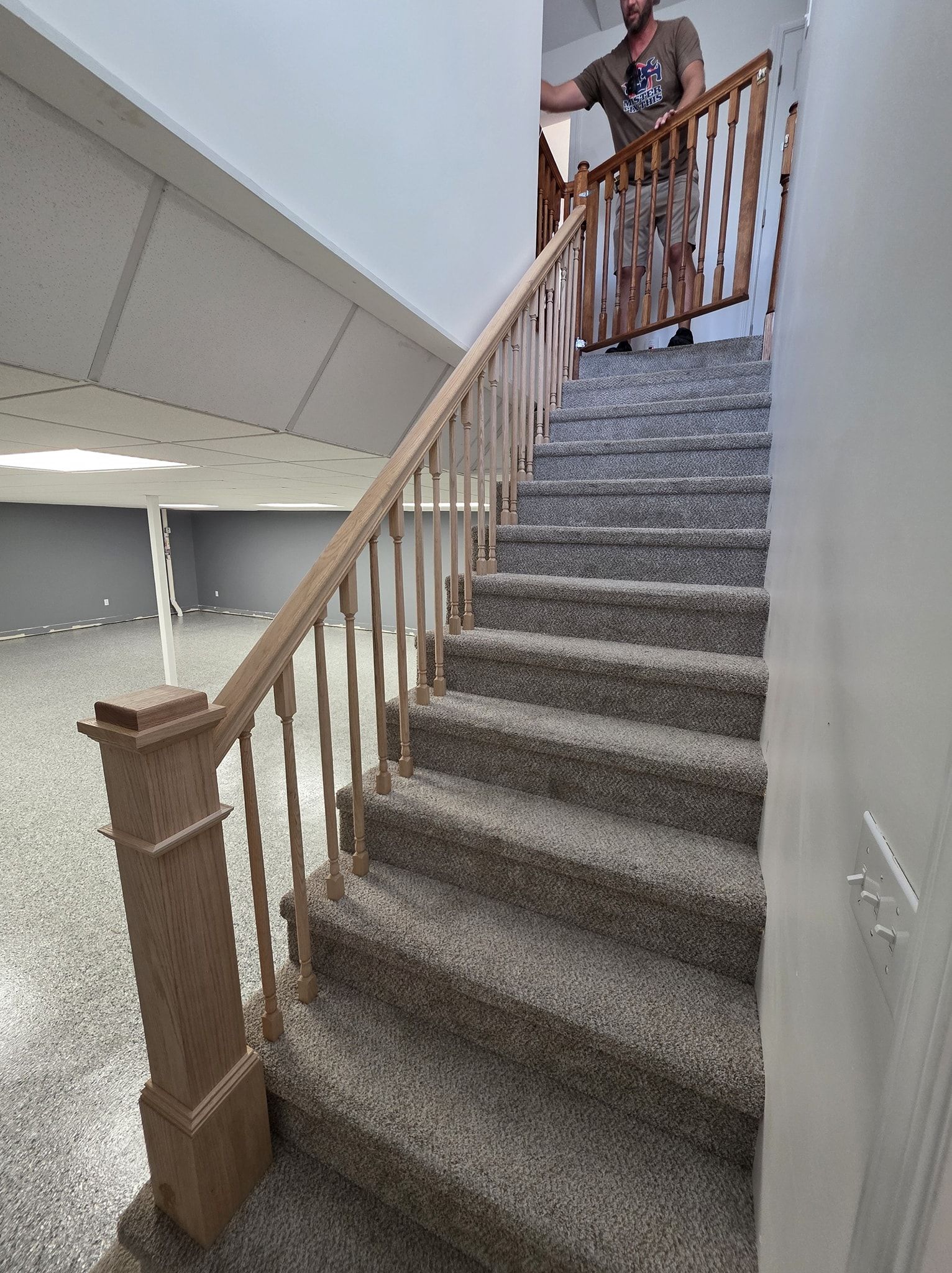 Person carrying a wooden stair railing up a carpeted staircase. Basement with gray walls and flooring.