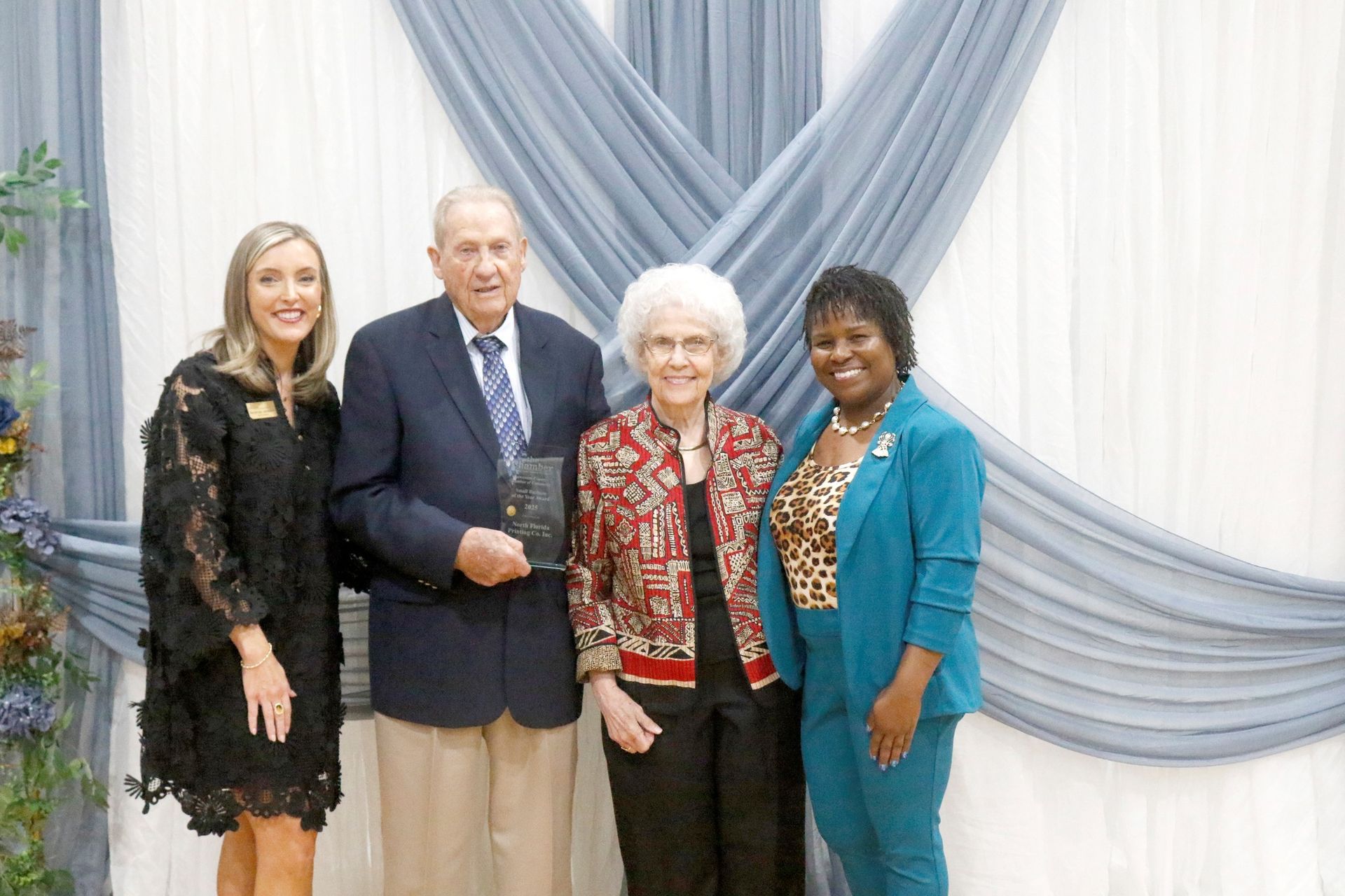 Four people stand together; a man in blue blazer, two women, and another woman in floral dress.