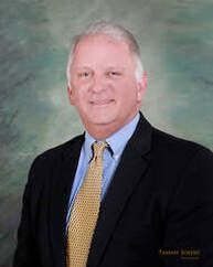 Man in a suit smiles, gold tie, blue shirt, dark jacket, against a mottled green-blue background.
