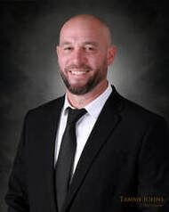 Man with shaved head and beard in a black suit, smiling at the camera.