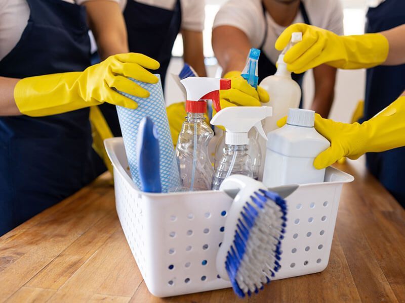 Hands in yellow gloves organize cleaning supplies in a white caddy on a wooden table.