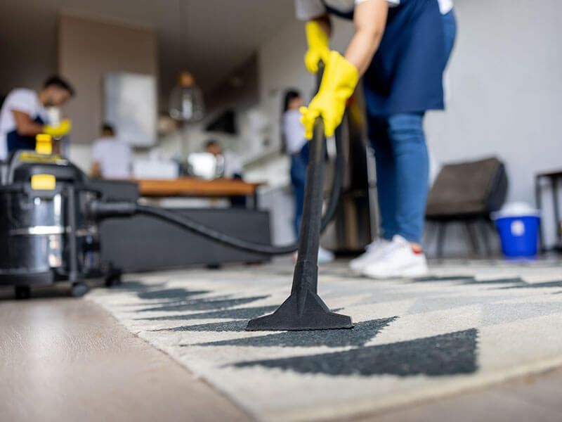 Person vacuuming a patterned rug in a living room, wearing yellow gloves and blue clothing.