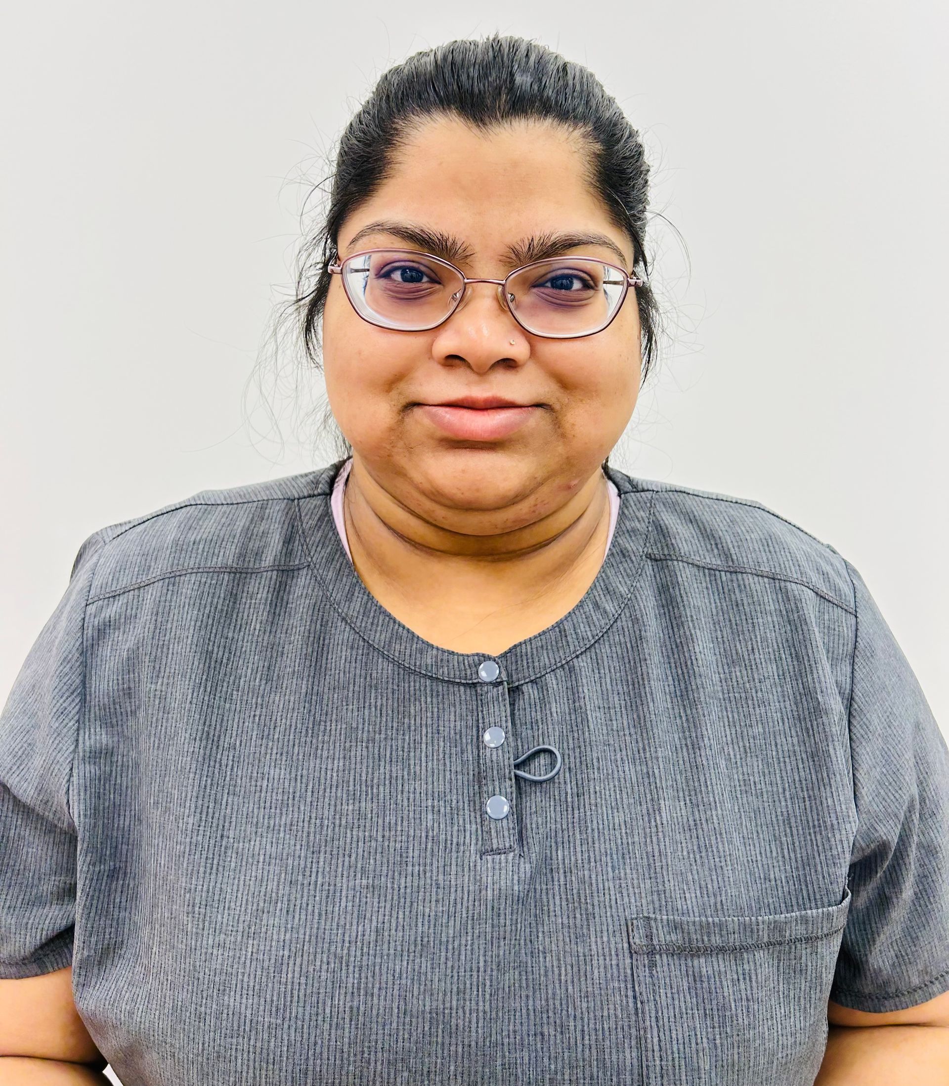 A woman wearing glasses and a grey shirt is standing in front of a white wall.