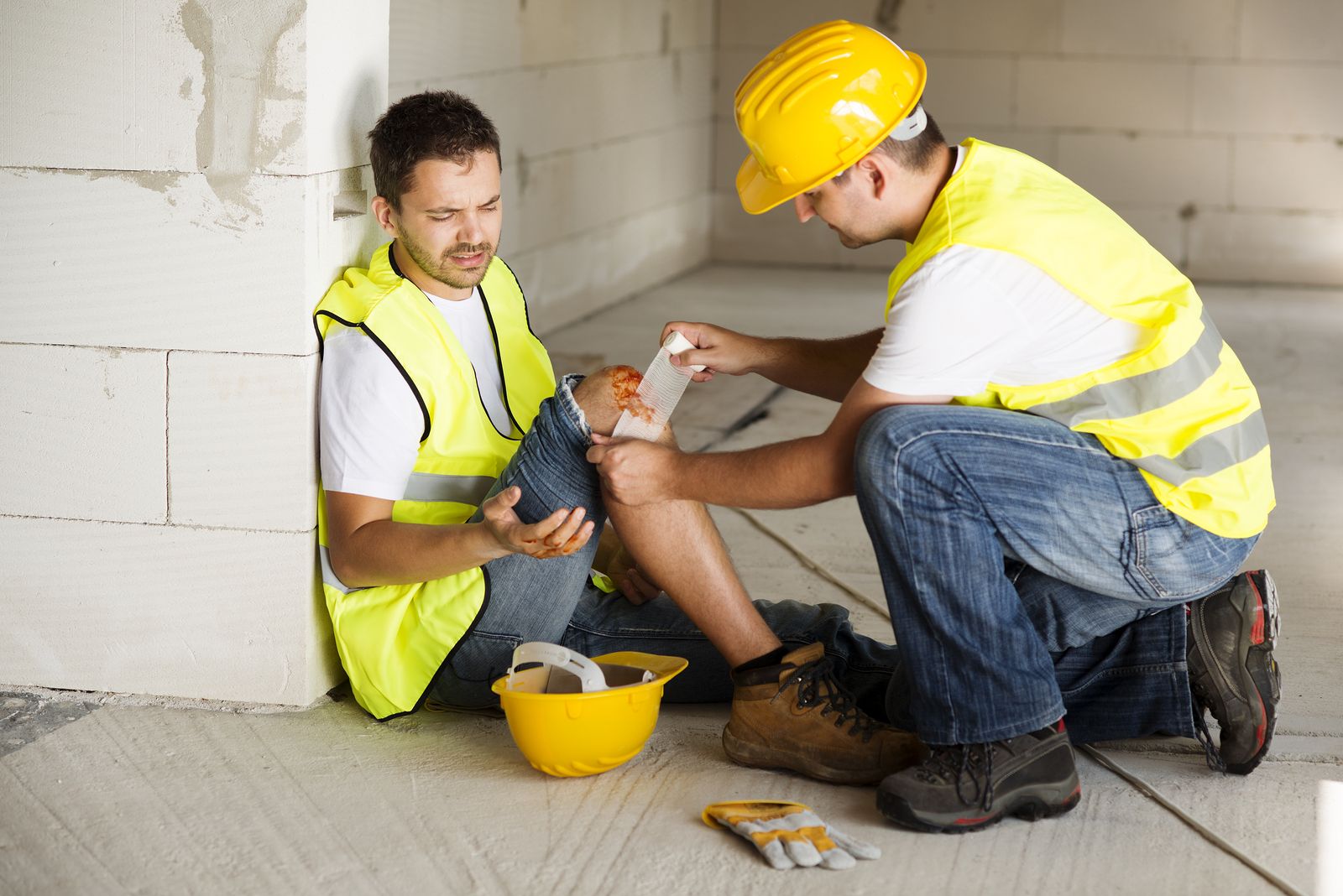 A construction worker is kneeling down next to another construction worker who has a knee injury.