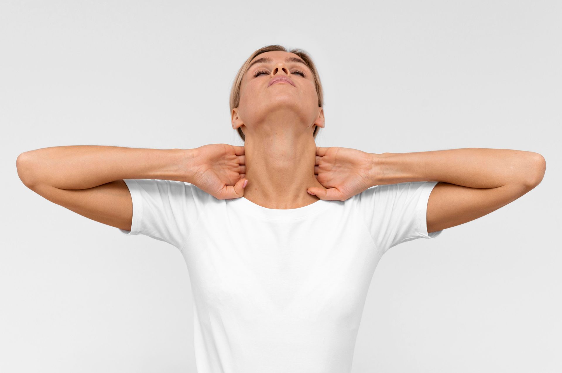 Woman in a white shirt stretching her neck with both hands against a plain background