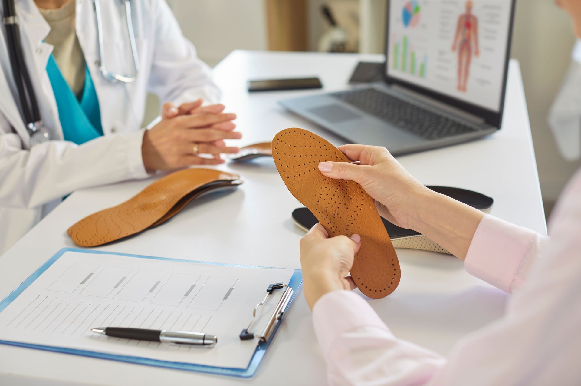Podiatrist discussing custom orthotic insoles with a patient at a desk with laptop and clipboard