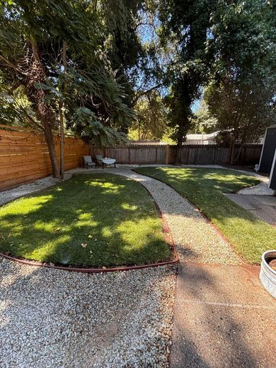 Sunlit backyard with curved gravel path, green lawn, wooden fence, and large trees shading the yard