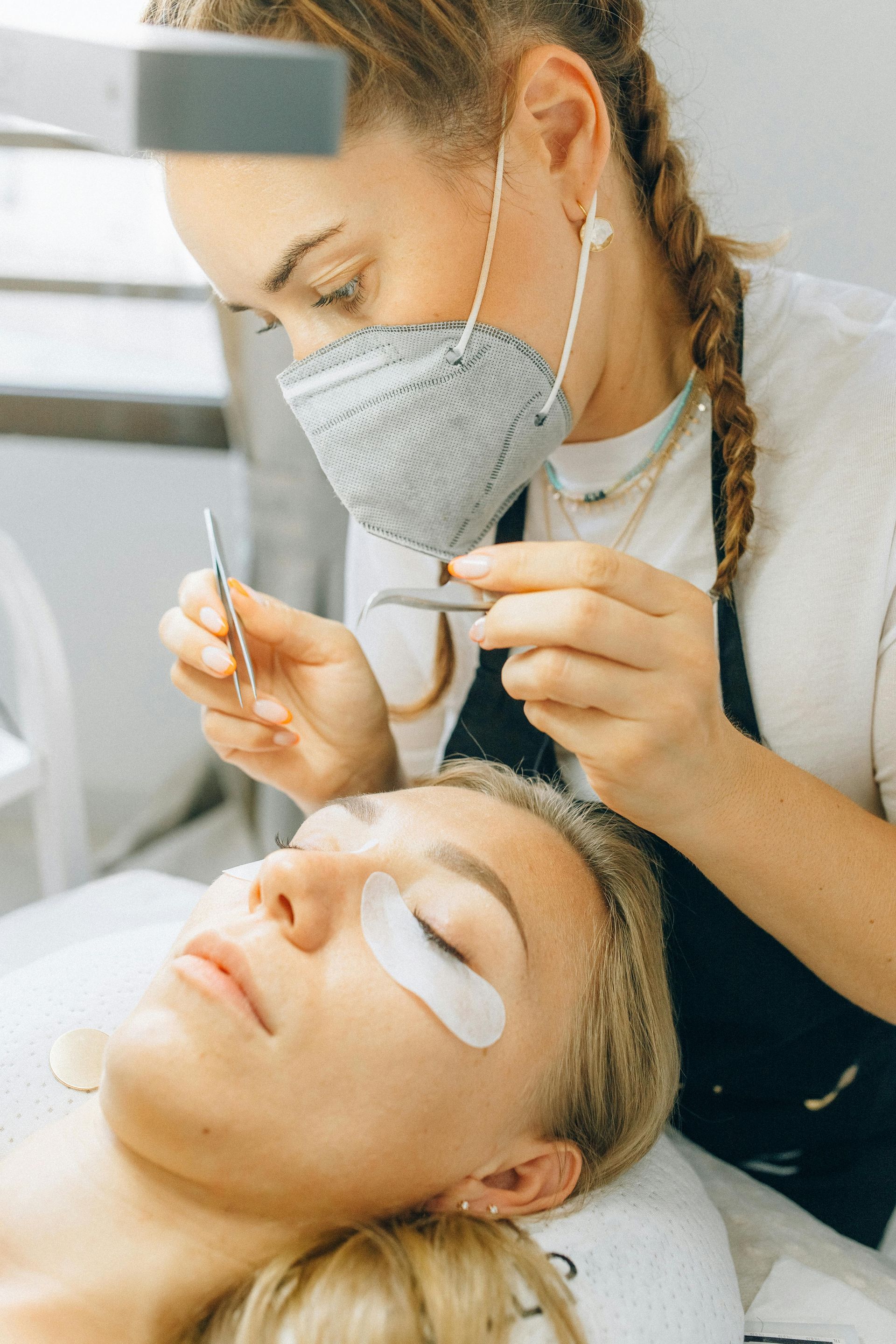 A woman wearing a mask is applying eyelash extensions to a woman.