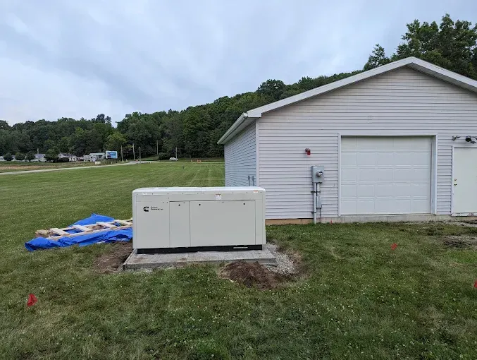 A white generator in front of a light-colored building with a garage door, on a grassy lawn with trees in the background.