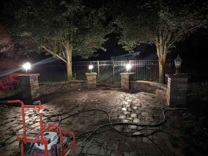 Night view of a brick patio with decorative lights on pillars, a metal fence, and two trees in the background.