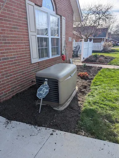 A home generator next to a brick house and flowerbed on a sunny day.