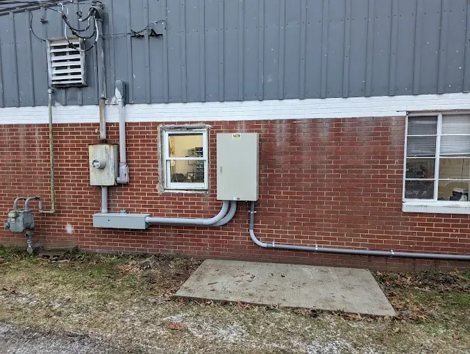 Exterior building wall with electrical boxes, conduits, and a concrete slab. Brick and gray siding.