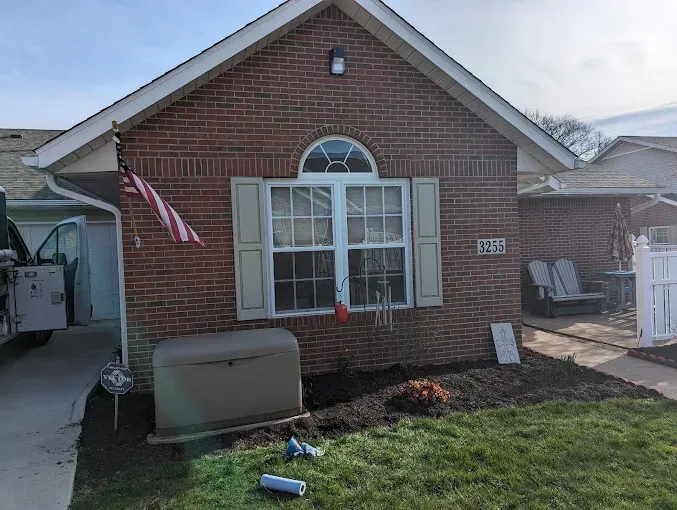 Brick house with a generator and American flag. Front yard with green grass, and garden bed.