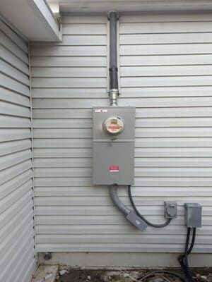 Gray electrical meter box on a light gray house wall with conduits and a service entrance.