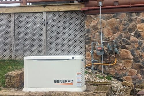 A beige Generac generator next to a gas meter on a stone wall, beside a wooden deck.
