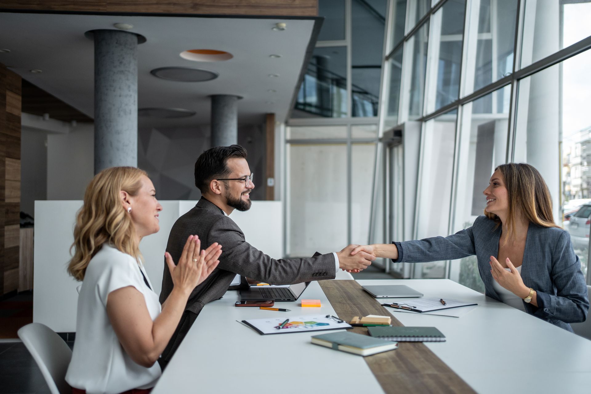 People shaking hands at a table in a modern office, one person clapping.
