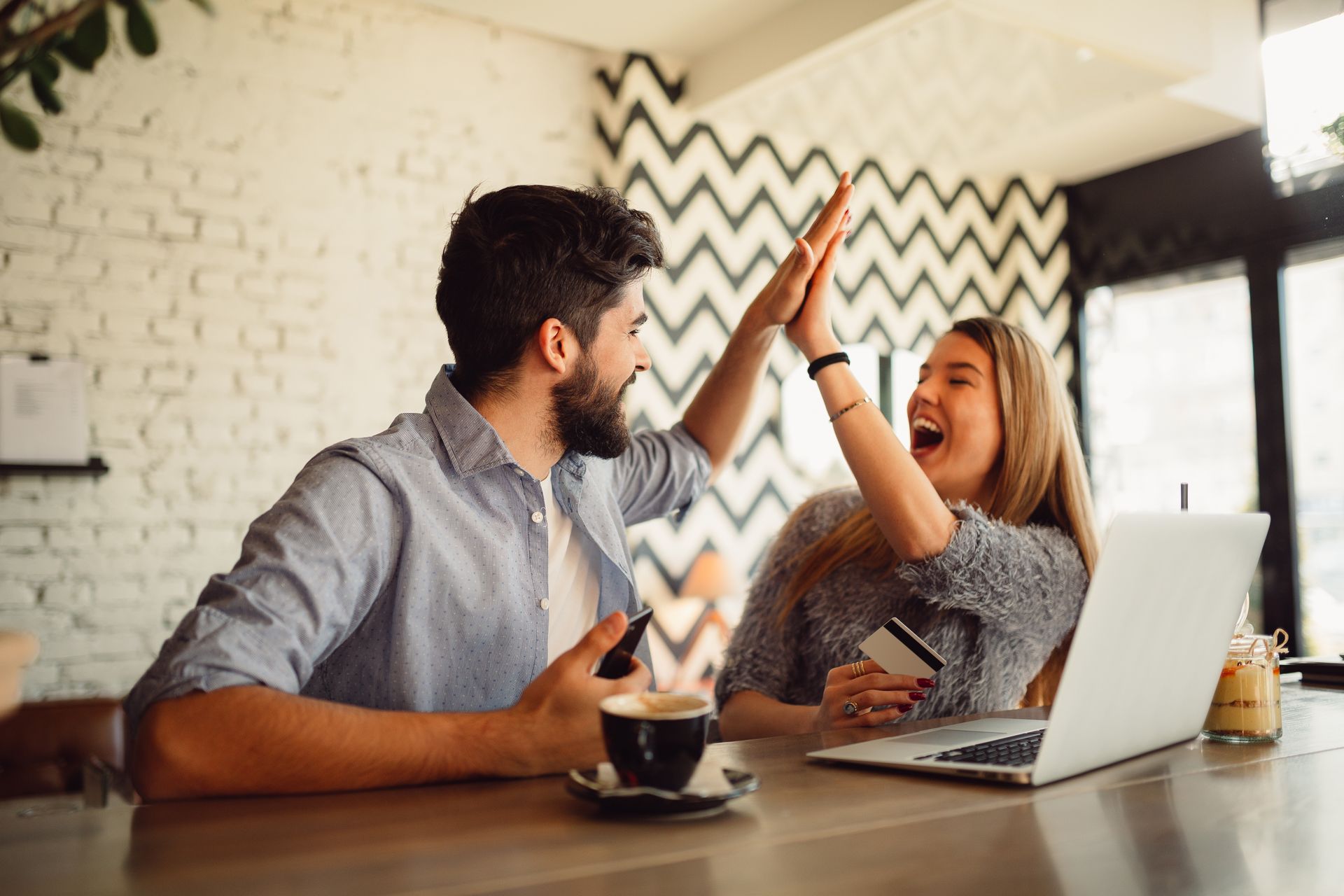 Couple high-fiving in cafe, celebrating success with laptop, coffee, and credit card.