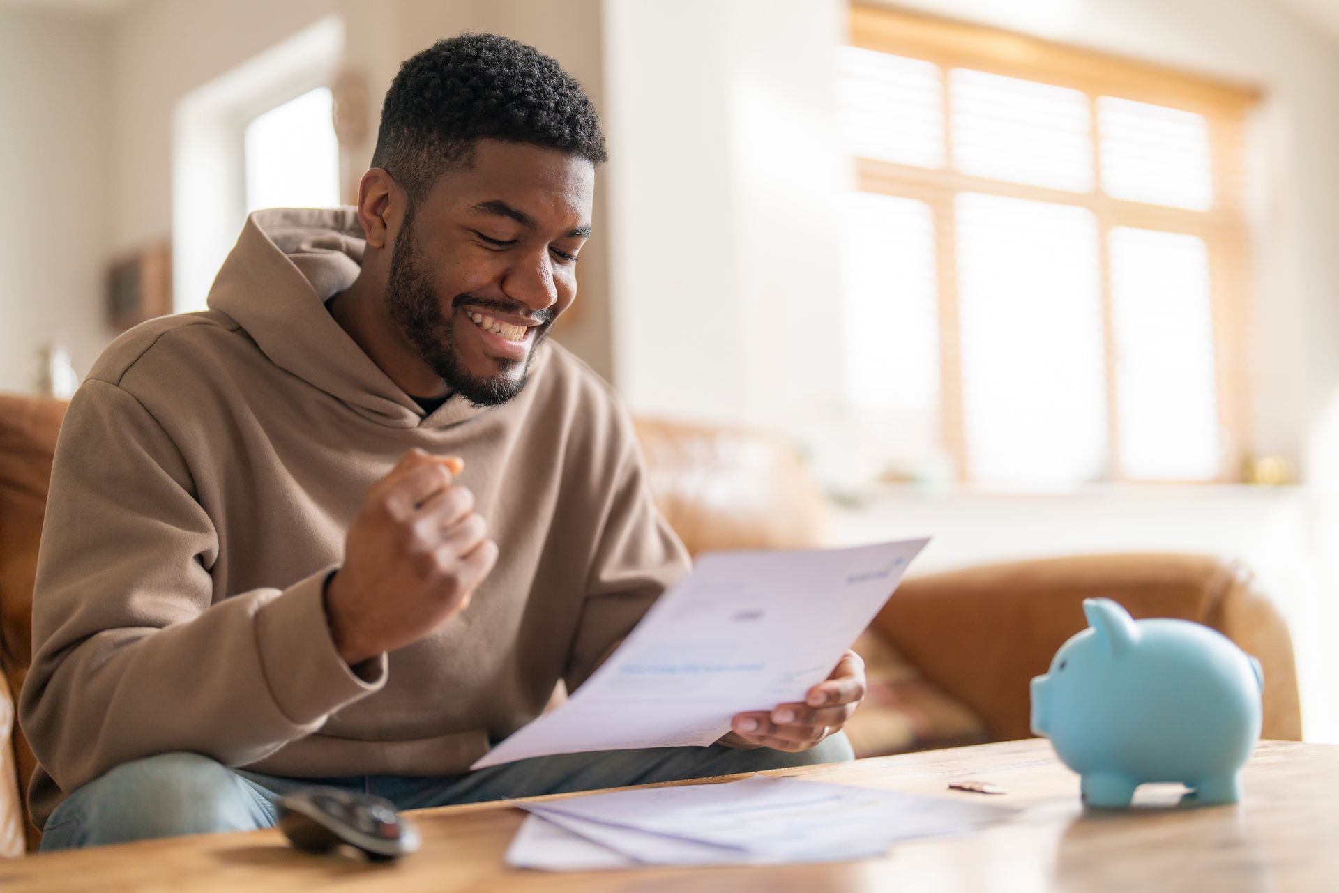 Man smiling, looking at paperwork with a piggy bank nearby; indoors, sunny.