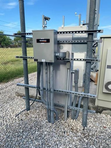 Electrical control panel with conduits mounted on a fenced utility site outdoors.