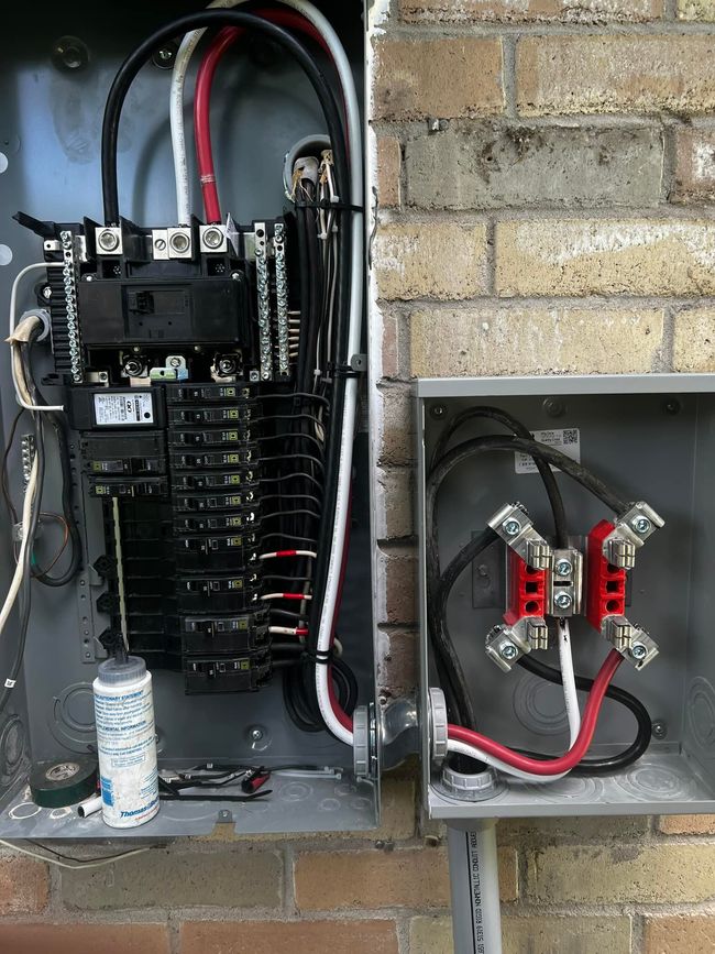 Open electrical panel with breakers and red cables mounted on a brick wall, with a smaller junction box beside it