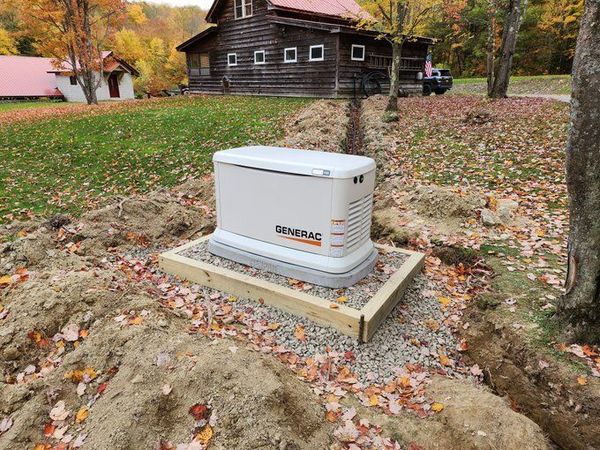 Outdoor Generac generator on a gravel pad beside a rustic cabin in autumn.
