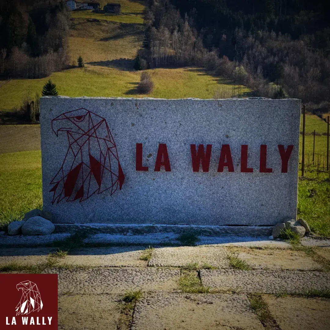 Ein großer, grauer Steinblock mit der roten Aufschrift „LA WALLY“ und einem geometrischen Adler-Logo in Rot steht auf einer gepflasterten Fläche vor einer grünen Wiese. Im Hintergrund erstrecken sich ein bewaldeter Hang und eine Berglandschaft mit einigen Häusern am oberen Bildrand.