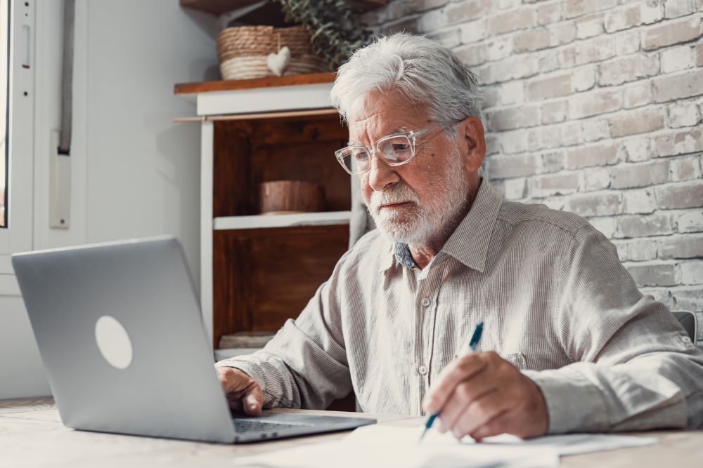An elderly man is sitting at a table using a laptop computer.