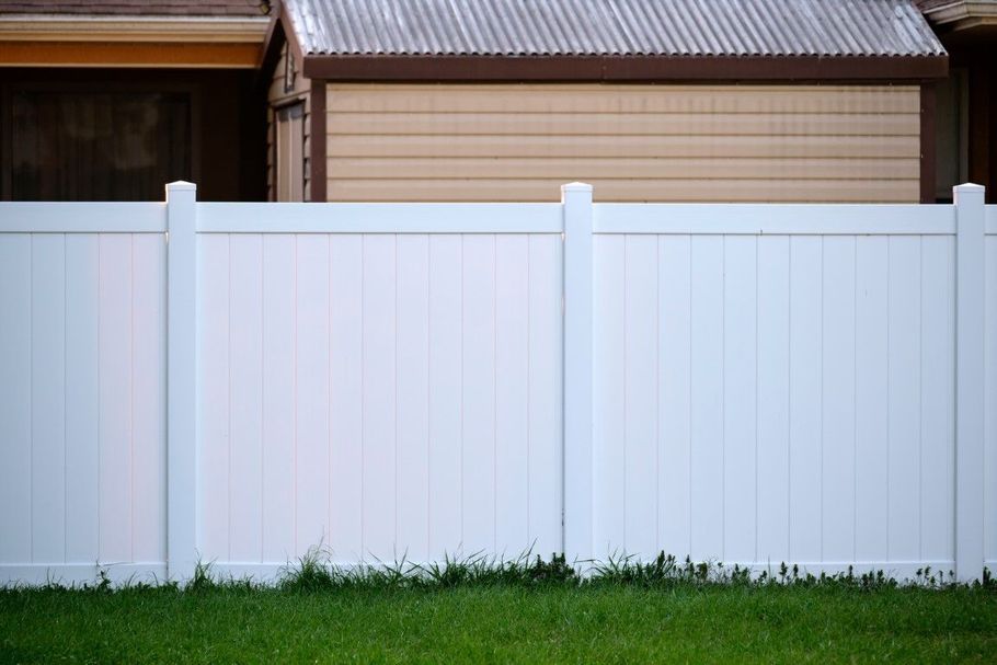 White vinyl fence in front of a beige house with a grass lawn underneath.
