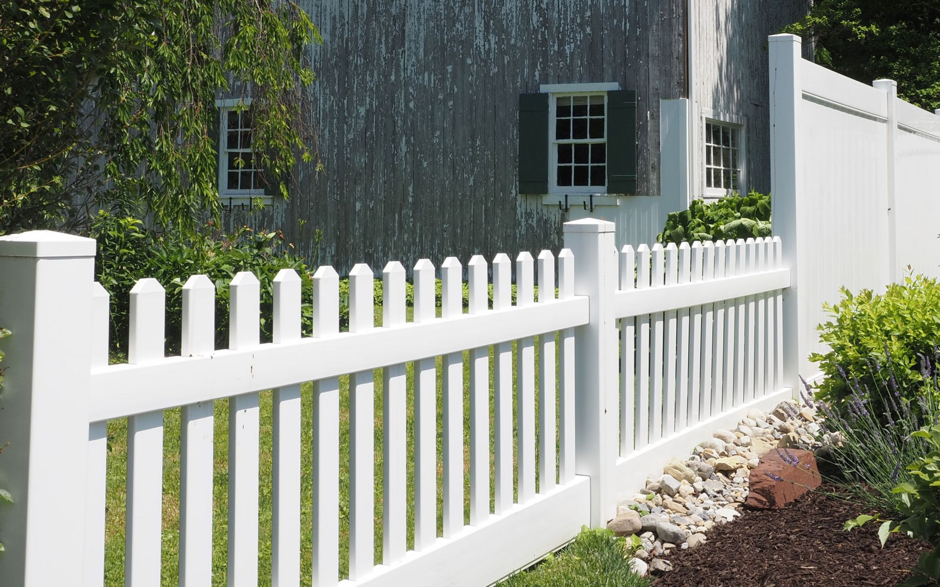 White picket fence along a house with green-trimmed windows and shrubs in the yard