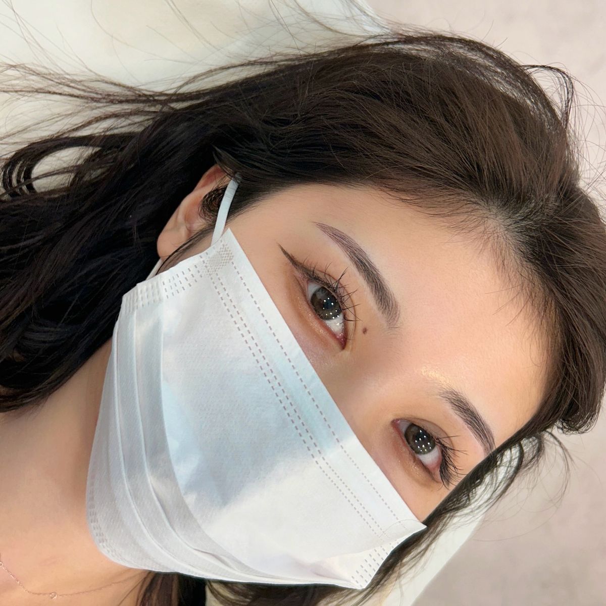 Woman wearing a surgical mask, dark hair framing face, light skin, and open eyes, lying down.