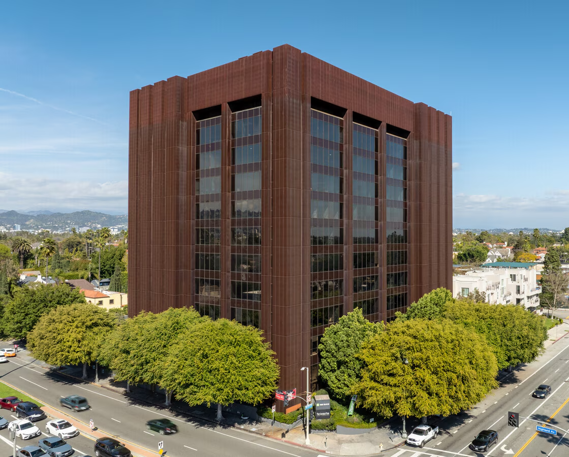 Tall rust-colored office building with reflective windows, seen from a street corner with trees in front.