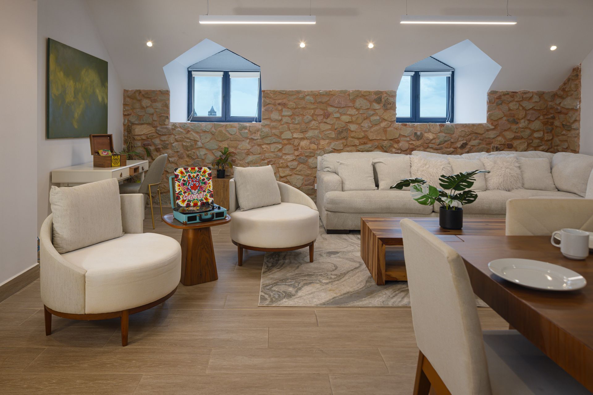 Living room at PH La Cuadra in Casco Viejo, with a stone accent wall, two recessed windows, cream armchairs, a sectional sofa, and wooden tables.