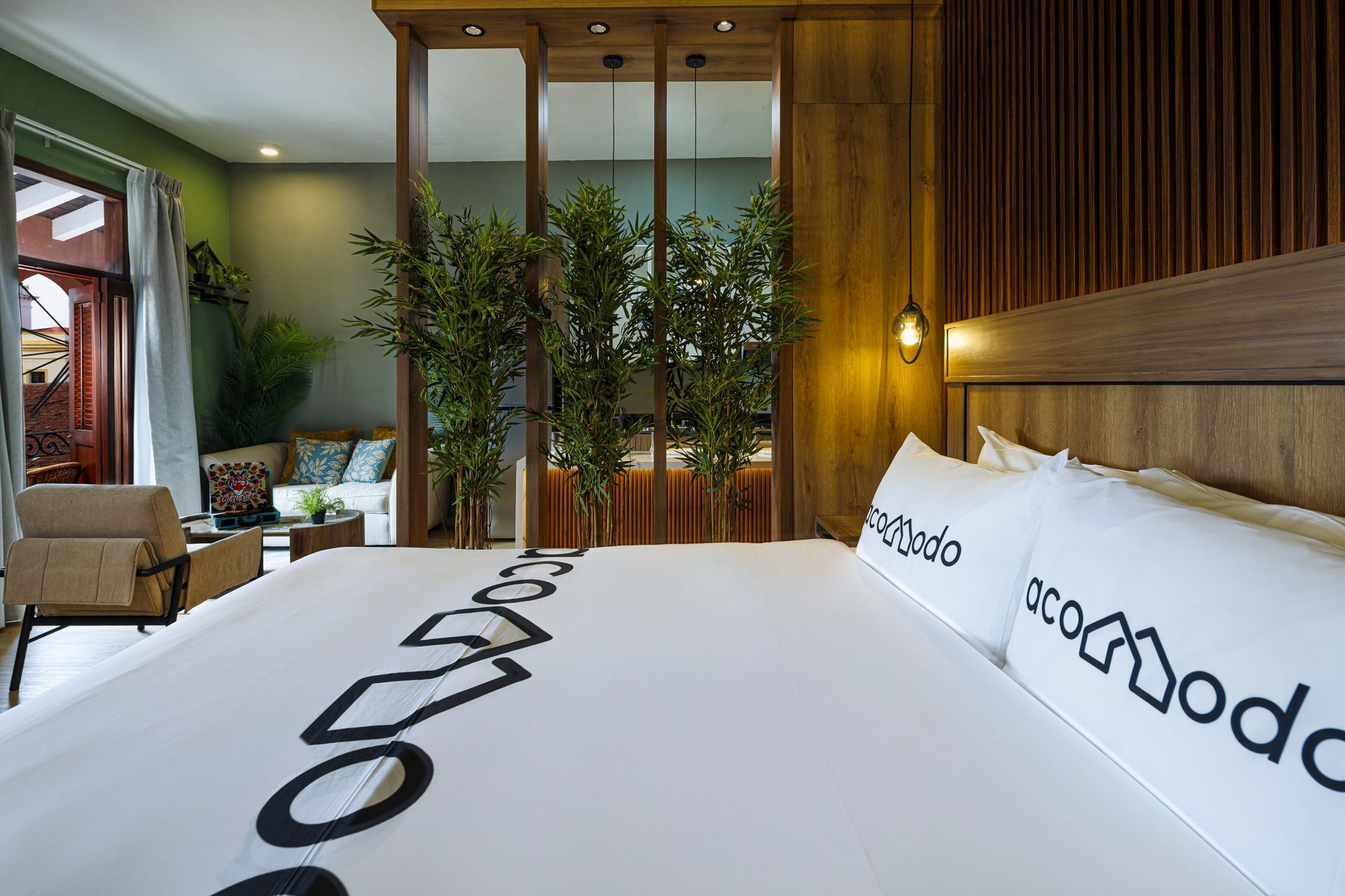 Bedroom at PH Hotel Colombia with white Acomodo bedding, a wooden headboard, warm bedside lighting, and plants separating the bed from the lounge.