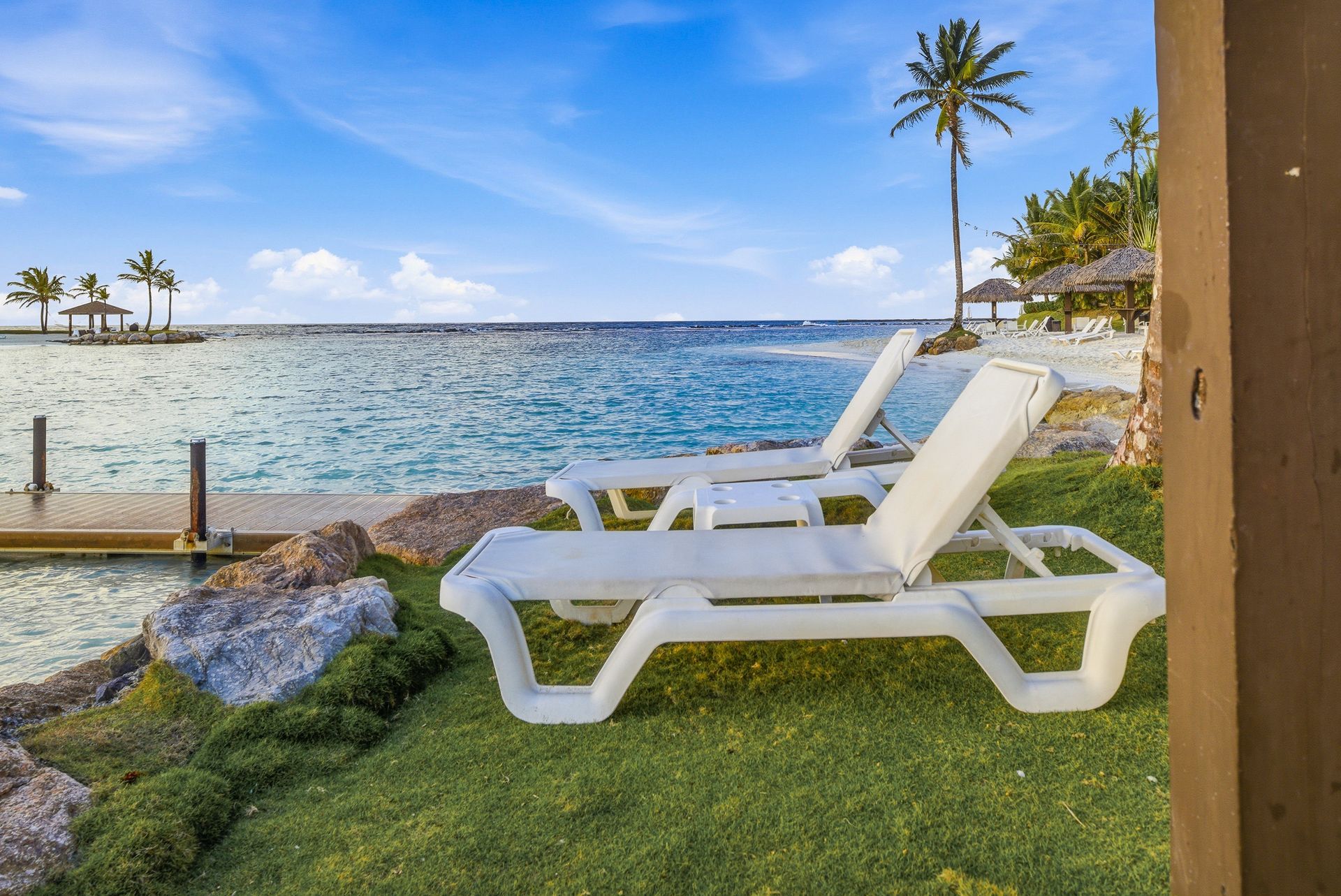 playa escondida colon White lounge chairs on a grassy shore overlooking a blue ocean with palm trees and a sunny sky 