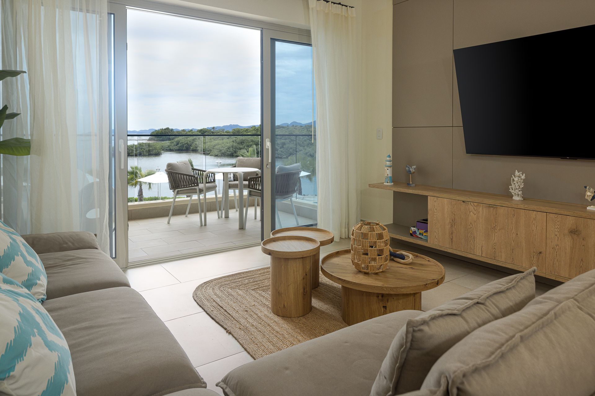 Bright living room with beige sofas, TV, woven tables, and sliding doors opening to a balcony and water view