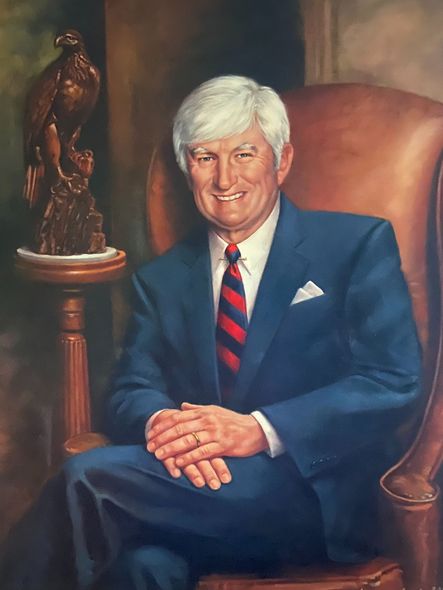 Portrait of a smiling man in a blue suit seated in a leather chair beside a bronze eagle statue.