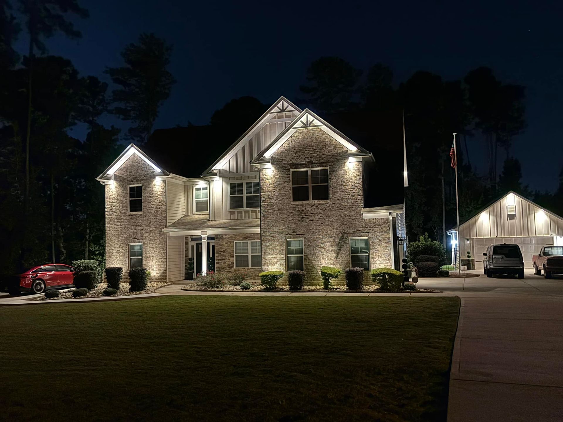 Two-story brick house illuminated at night. Features include garage, red car, and manicured lawn.