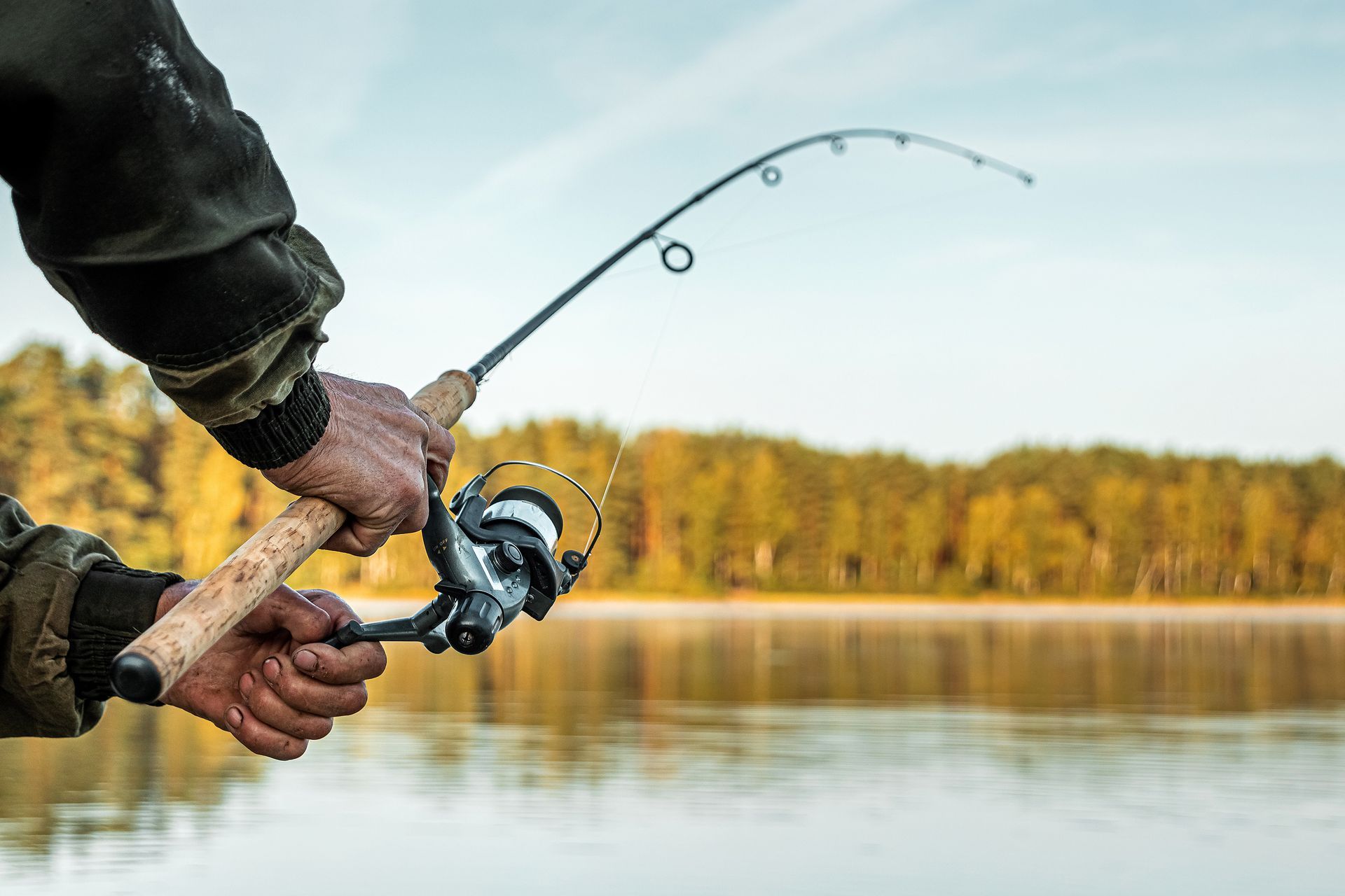 Person fishing with a rod and reel at a lake, holding the rod with both hands. Trees line the distant shore.