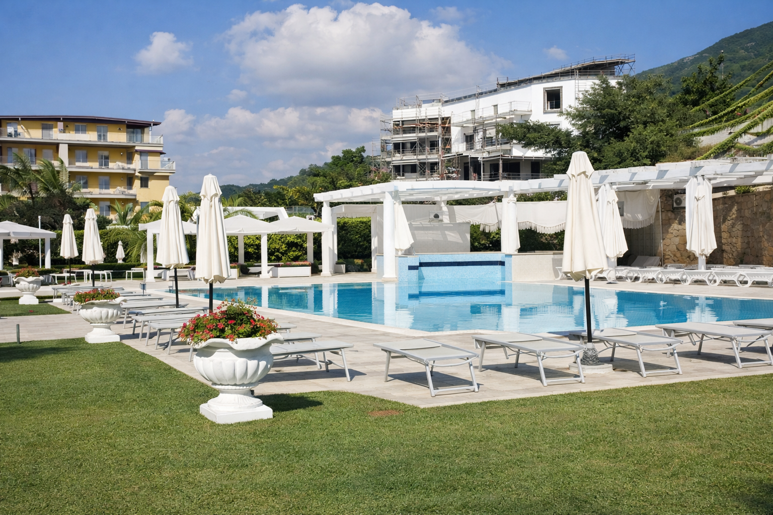 Piscina con sedie a sdraio, ombrelloni bianchi e strutture alberghiere sotto un cielo azzurro.