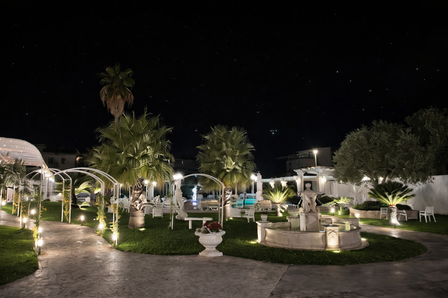 Giardino notturno con vialetti illuminati, alberi, fontane e strutture bianche.
