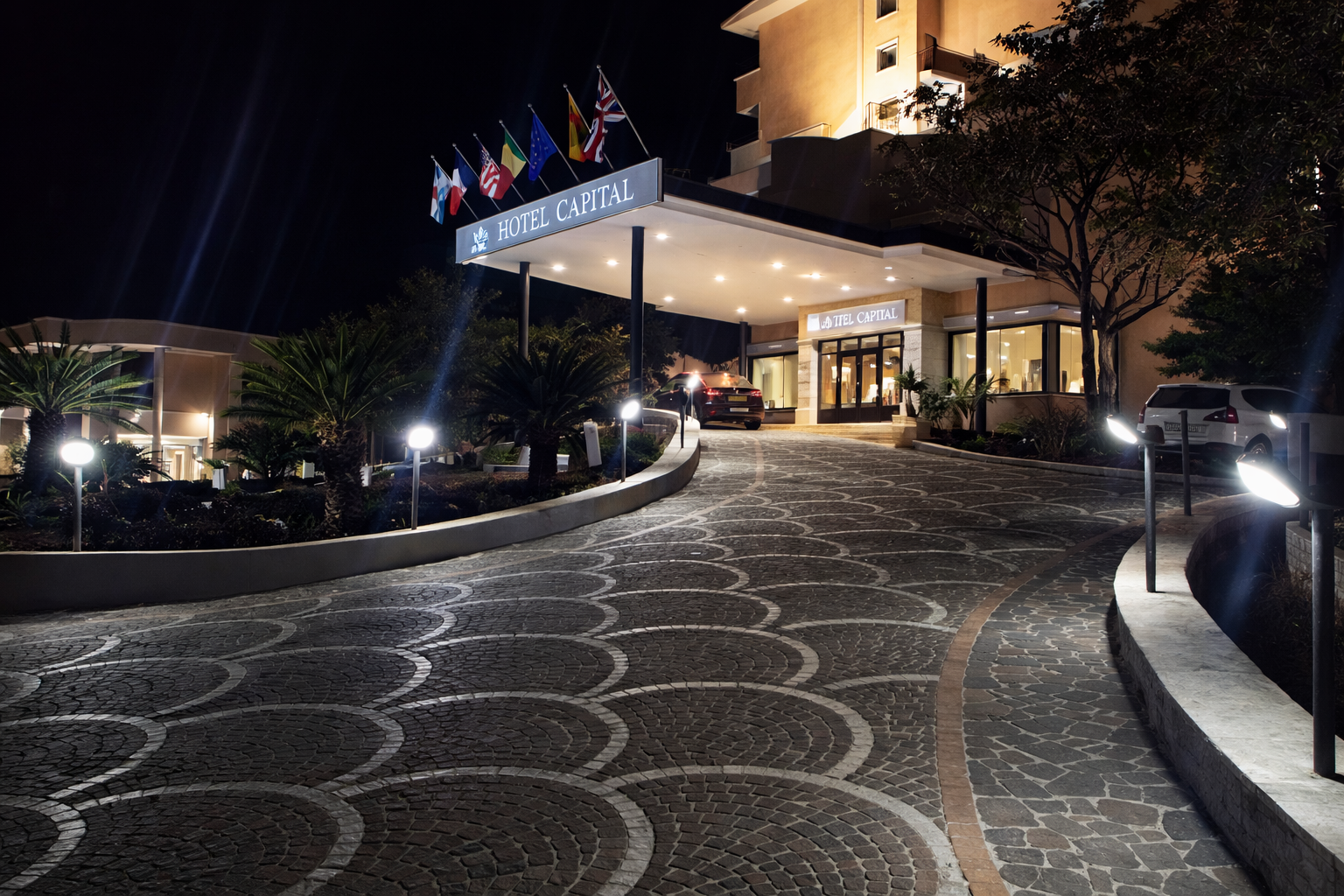 Ingresso dell'hotel di notte con passerella illuminata. Bandiere in cima all'ingresso, giardino su entrambi i lati.