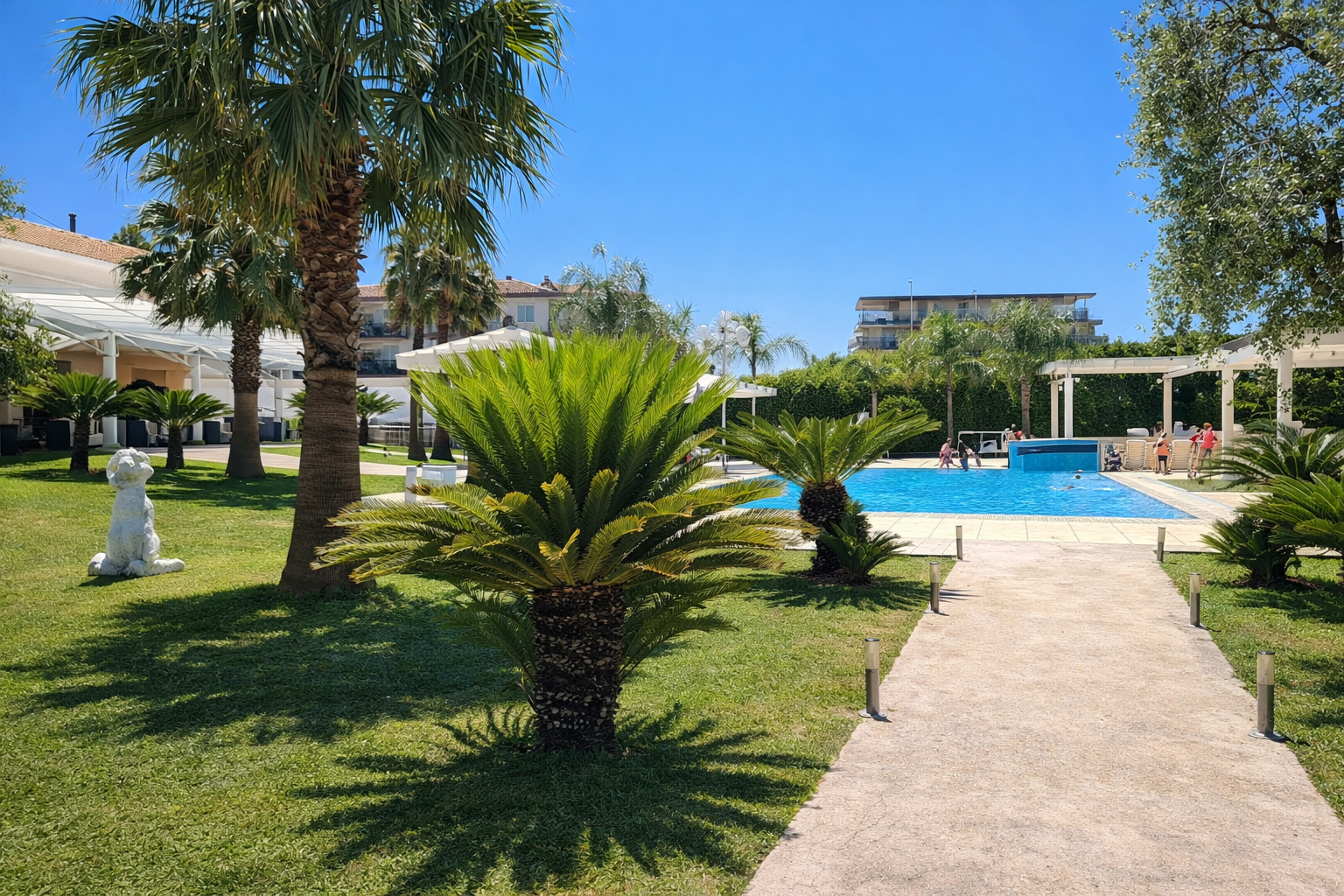 Un rigoglioso giardino verde con palme, una piscina e un sentiero sotto un cielo azzurro brillante.