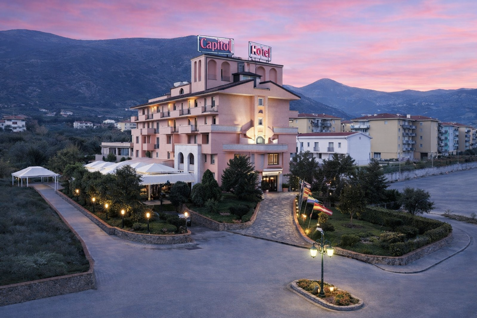 Edificio dell'hotel con insegna, sentiero d'ingresso, luci, alberi e montagne sullo sfondo al tramonto.